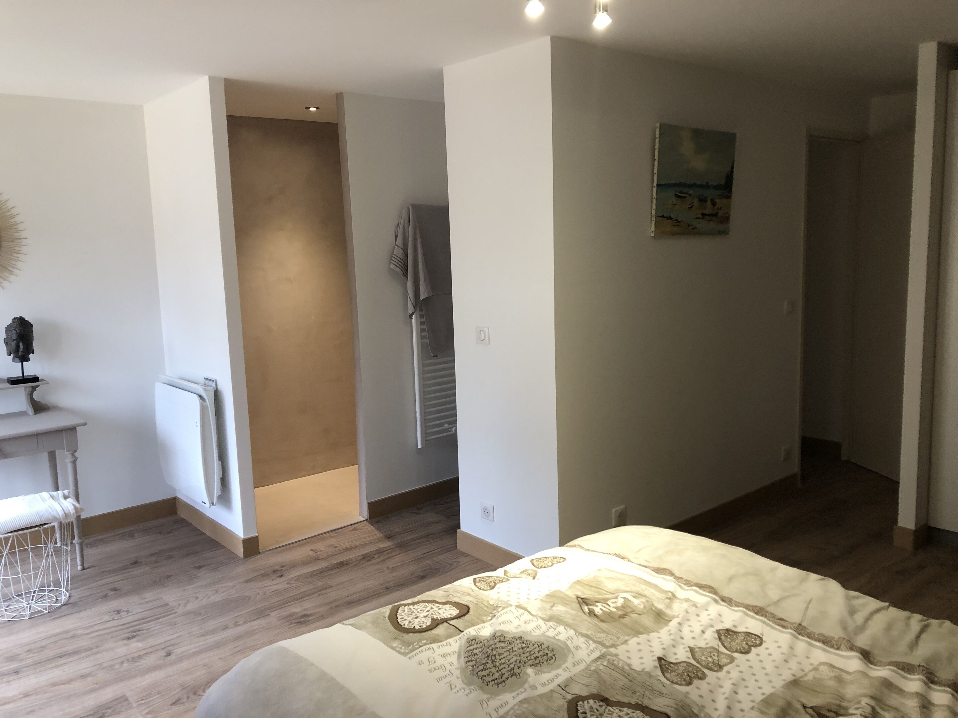 A bedroom with a wooden floor, a bed with a patterned bedspread, a small table with a chair, and a painting on the wall.