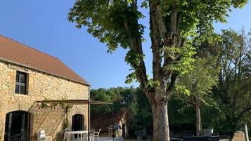 Un bâtiment en pierre avec un toit en tuiles, un patio couvert avec des chaises et un grand arbre au premier plan.