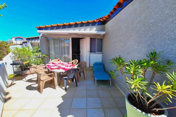 A small patio with a table set for two, a blue lounge chair, and potted plants.