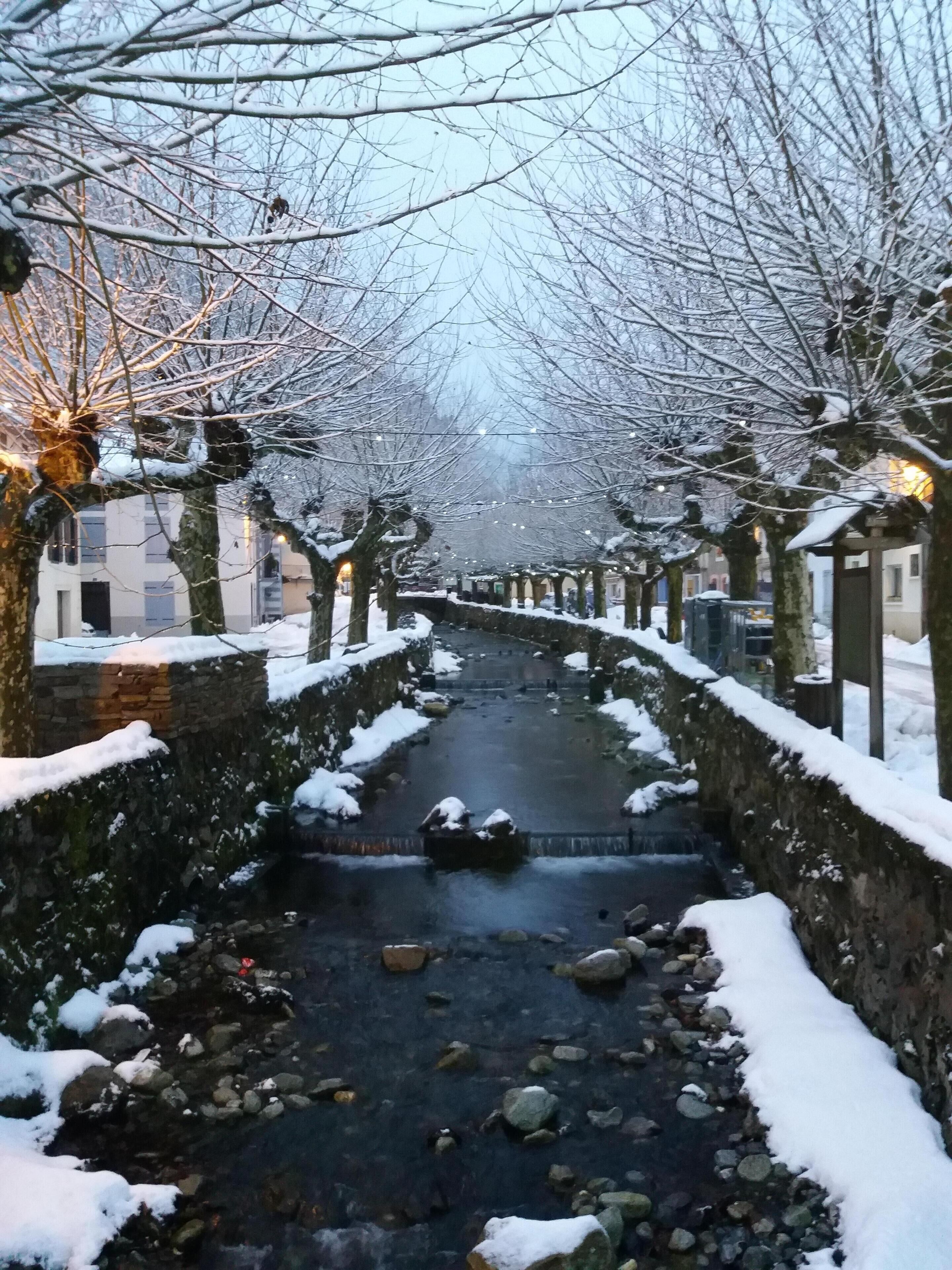 Un sendero cubierto de nieve, bordeado por muros de piedra y árboles desnudos, que lleva a un pequeño puente sobre un arroyo.