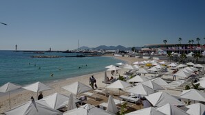 Una playa con sombrillas blancas, un muelle y barcos al fondo.
