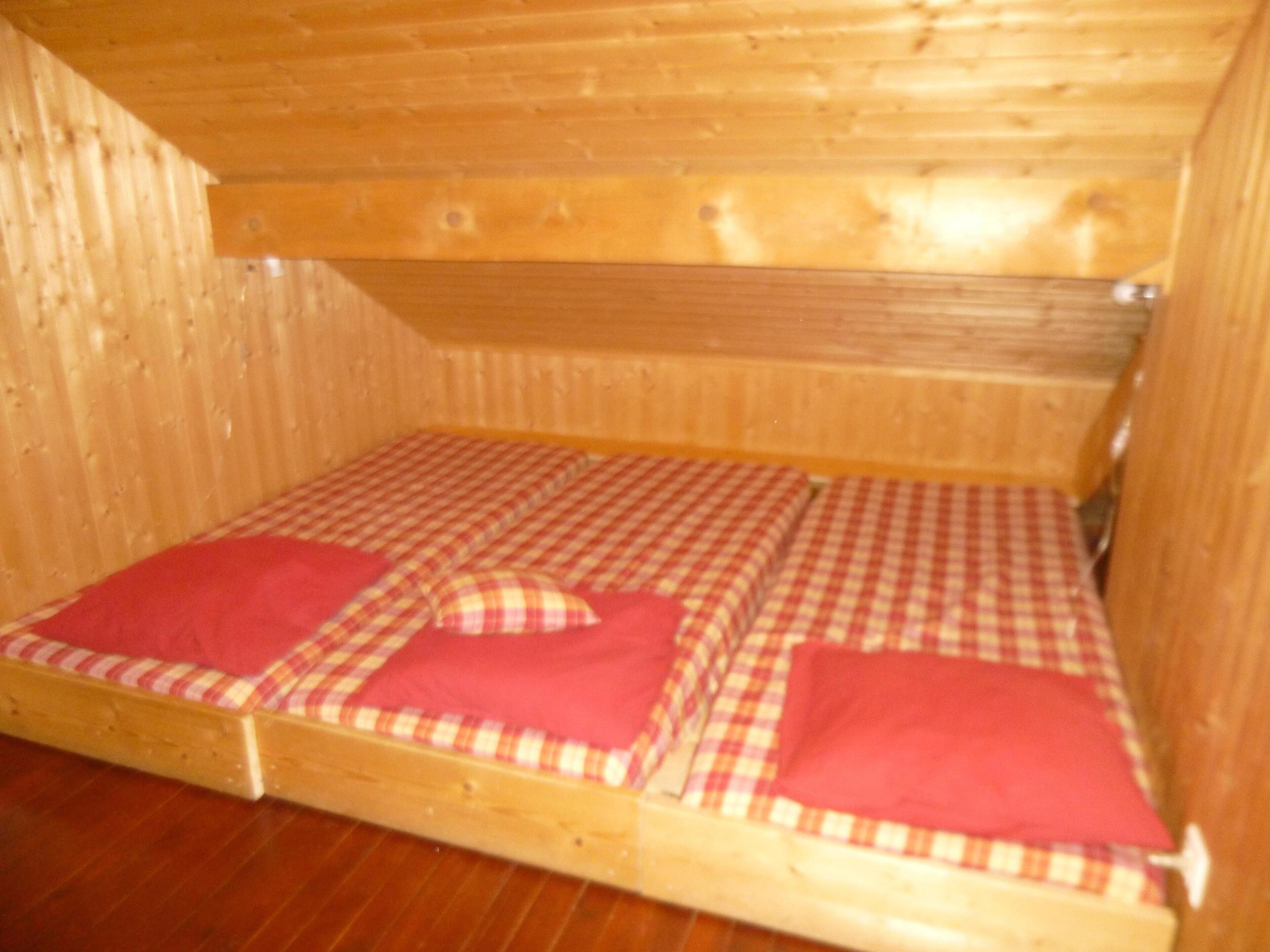 Une petite cabane en bois avec un lit simple, doté d’une literie à carreaux et de coussins rouges.