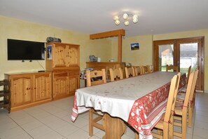A dining area with a long wooden table, chairs, a television mounted on the wall, and a kitchen area with wooden cabinets and a sink.
