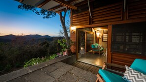 A wooden cabin with a stone patio, a green cushioned outdoor seating area, and a view of the mountains.