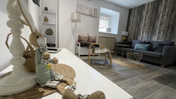 En moderne stue med en hvid sofa, en grå sofa, en træstol og et hvidt sofabord. Der er en lampe, en reol med pynteting og et vindue med gardiner.