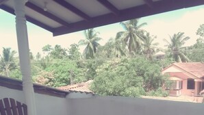 A covered balcony with wooden furniture, a small table with a flower, and a view of palm trees and a house in the background.
