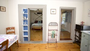 A room with a bed, a wooden floor, a small table with a tablecloth, a chair, a shelf with decorative items, and a microwave on a cabinet. - Martha's Vineyard 1BR, Pets Ok, 10 Min walk to beach or Edgartowns shops! (Edgartown)
