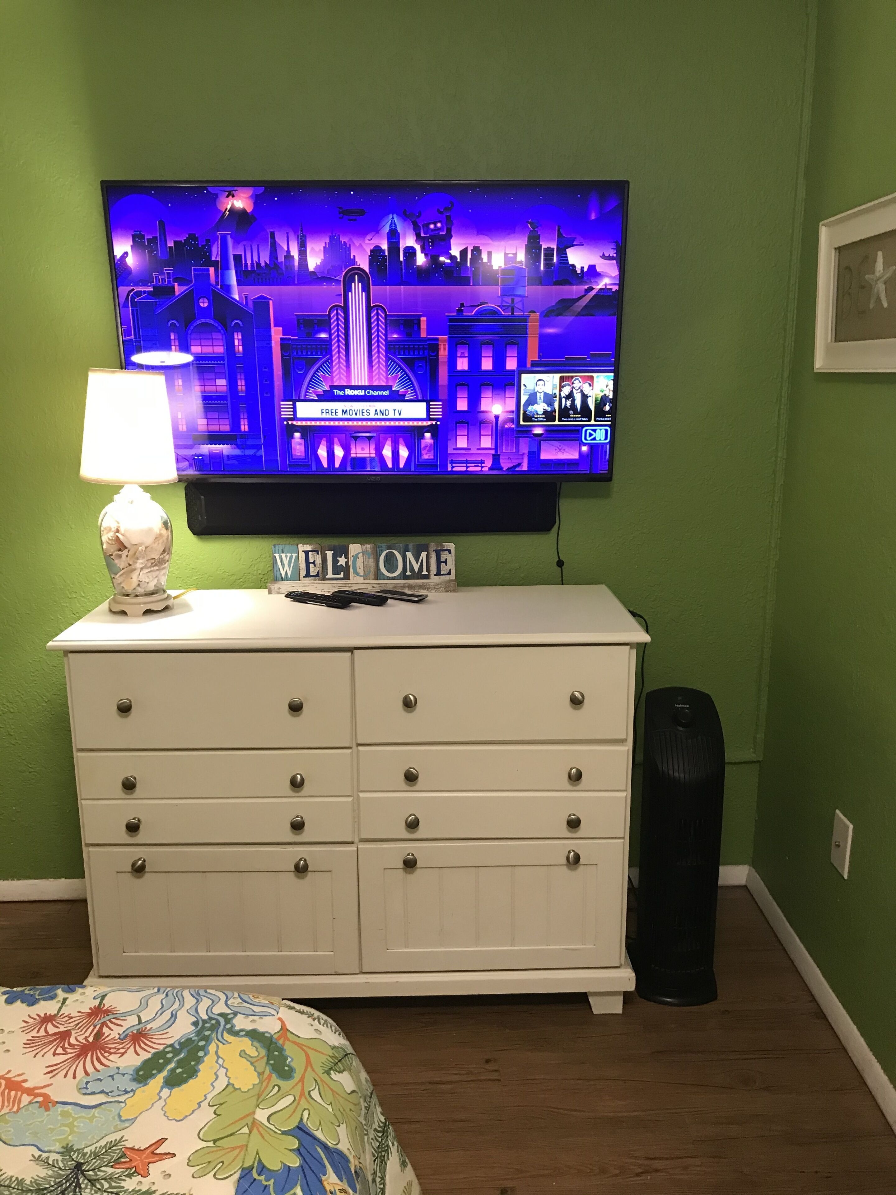 A room with a television displaying a cityscape, a white dresser with drawers, a lamp, and a welcome sign.