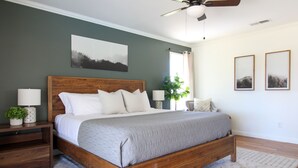 Een slaapkamer met een houten bed, een nachtkastje met een lamp, een plafondventilator en een schilderij aan de muur.