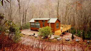 A cabin with a deck, surrounded by trees and a rocky ground.