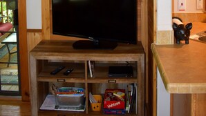 A wooden entertainment center with a flat-screen TV, shelves with books and remotes, a clock on the wall, and a small black figurine on the counter.