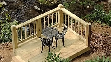 A wooden deck with a table and chairs, surrounded by greenery and a rocky stream.