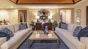 Ein großzügiges Wohnzimmer mit hoher Decke, einem zentralen Couchtisch, zwei Sofas mit gemusterten Kissen, einem dekorativen Spiegel und einer Topfpflanze.