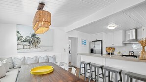 A modern kitchen with white cabinets, a stainless steel refrigerator, and a gas stove. There is a wooden dining table with a yellow tray and white chairs.