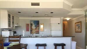 Een moderne keuken met witte kasten, een granieten aanrecht en een centraal eiland met krukken. Twee overheadlampen en een ventilatieopening zijn in het plafond aangebracht.