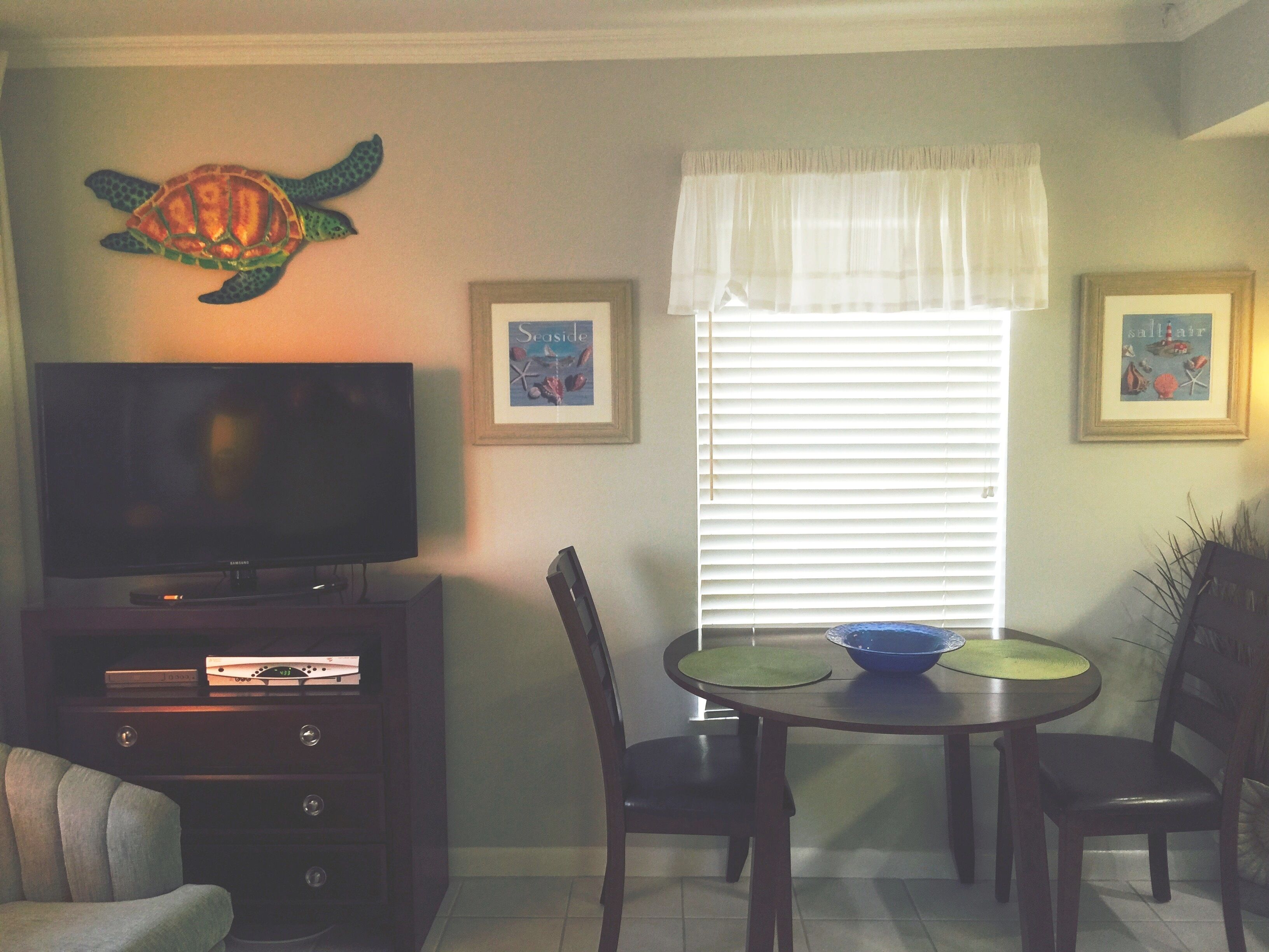 A living room with a flat-screen TV, a dining table with chairs, a wall-mounted turtle decoration, and framed artwork.