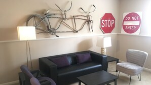 Een woonkamer met een zwarte bank, paarse fauteuils, een salontafel, een lamp en een aan de muur bevestigd fietsbeeldhouwwerk.