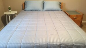 Een bed met een geborduurde blauwe spreideken, een klein rond tafeltje met een cactus, en een houten nachtkastje met een lade.