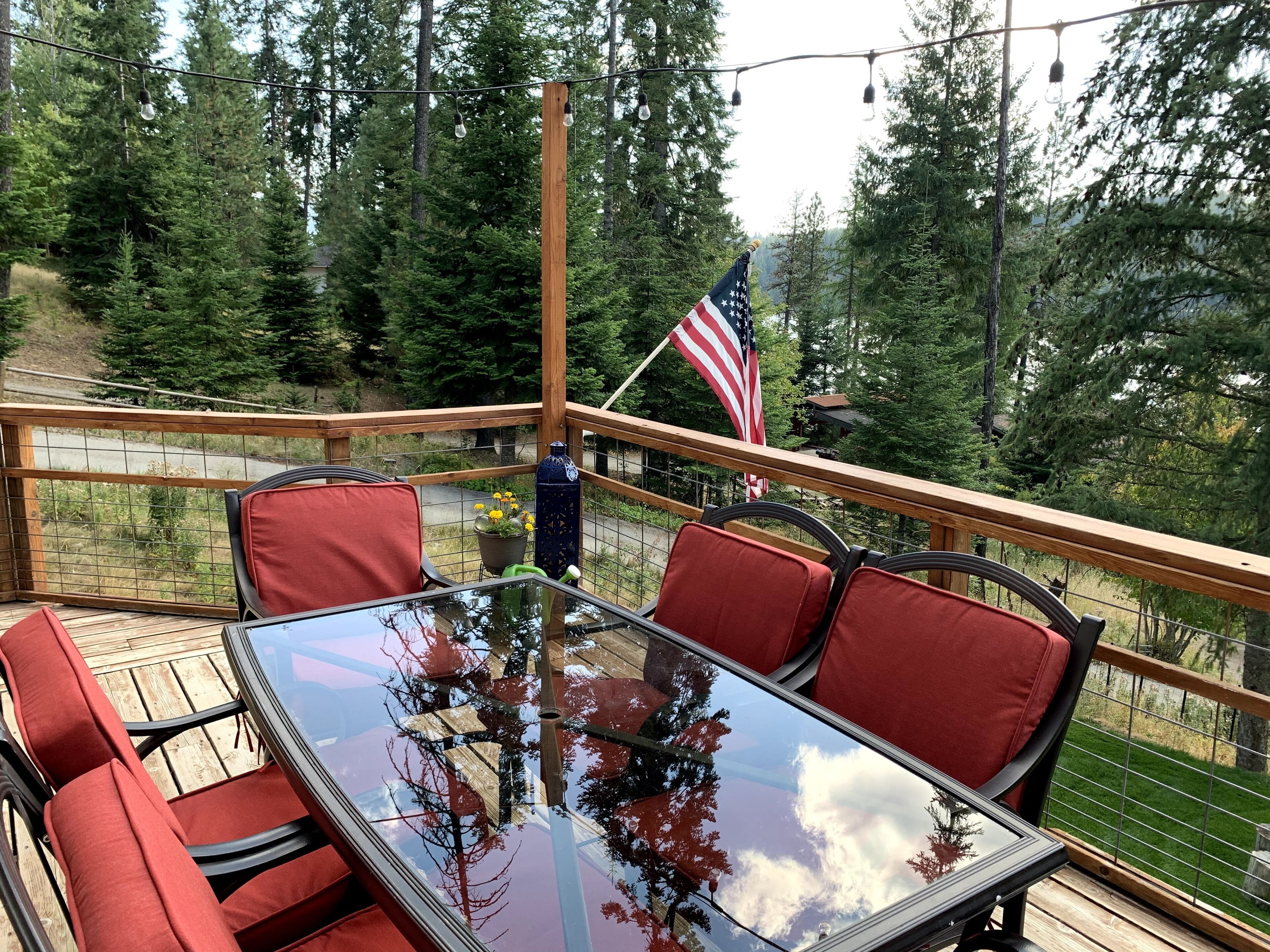 A deck with a glass-top table and red cushioned chairs, surrounded by a wooden railing, with an American flag and string lights.