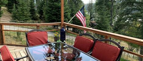 A deck with a glass-top table and red cushioned chairs, surrounded by a wooden railing, with an American flag and string lights.