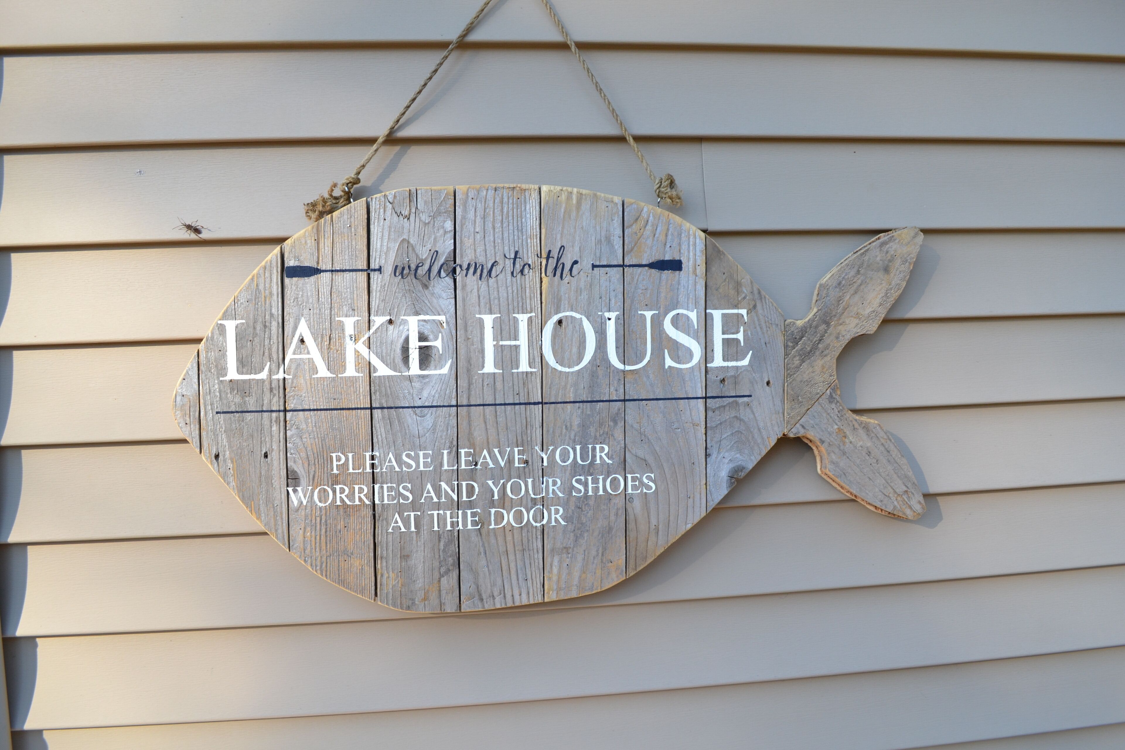 Une pancarte en bois portant l’inscription « LAKE HOUSE » et le message : « VEUILLEZ LAISSER VOS SOUCIS ET VOS CHAUSSURES À L’ENTRÉE. »