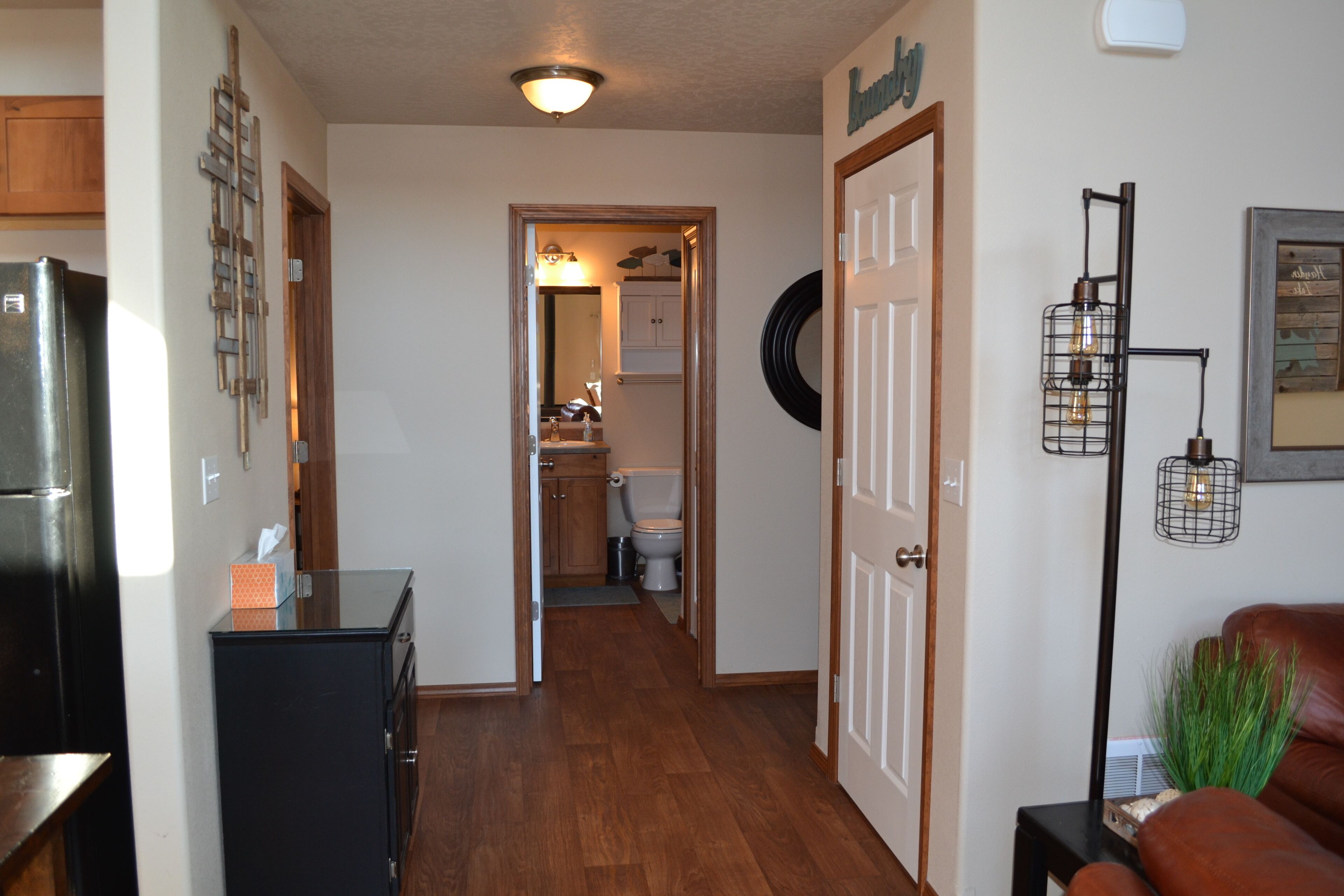 A hallway with a refrigerator, a cabinet, a door with a nameplate, a wall-mounted light fixture, and a plant on a side table.