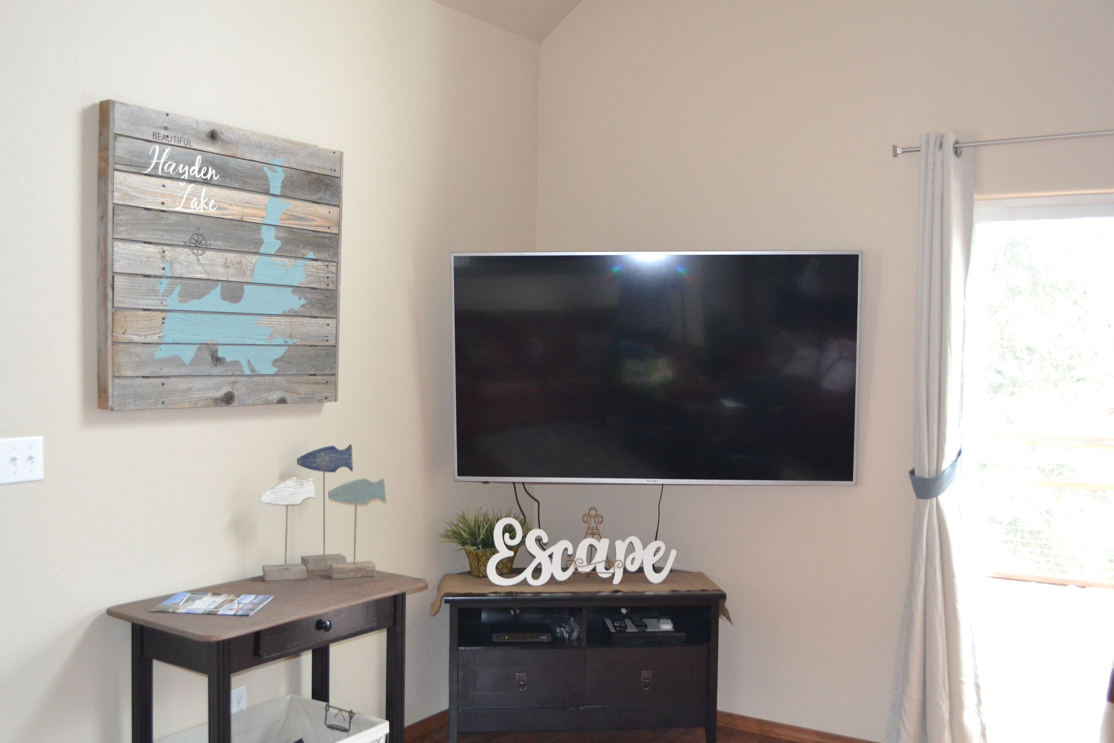 Una habitación con un televisor de pantalla plana montado en la pared, un letrero de madera con texto, una mesita con adornos y un mueble debajo del televisor.