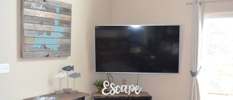 Una habitación con un televisor de pantalla plana montado en la pared, un letrero de madera con texto, una mesita con adornos y un mueble debajo del televisor.