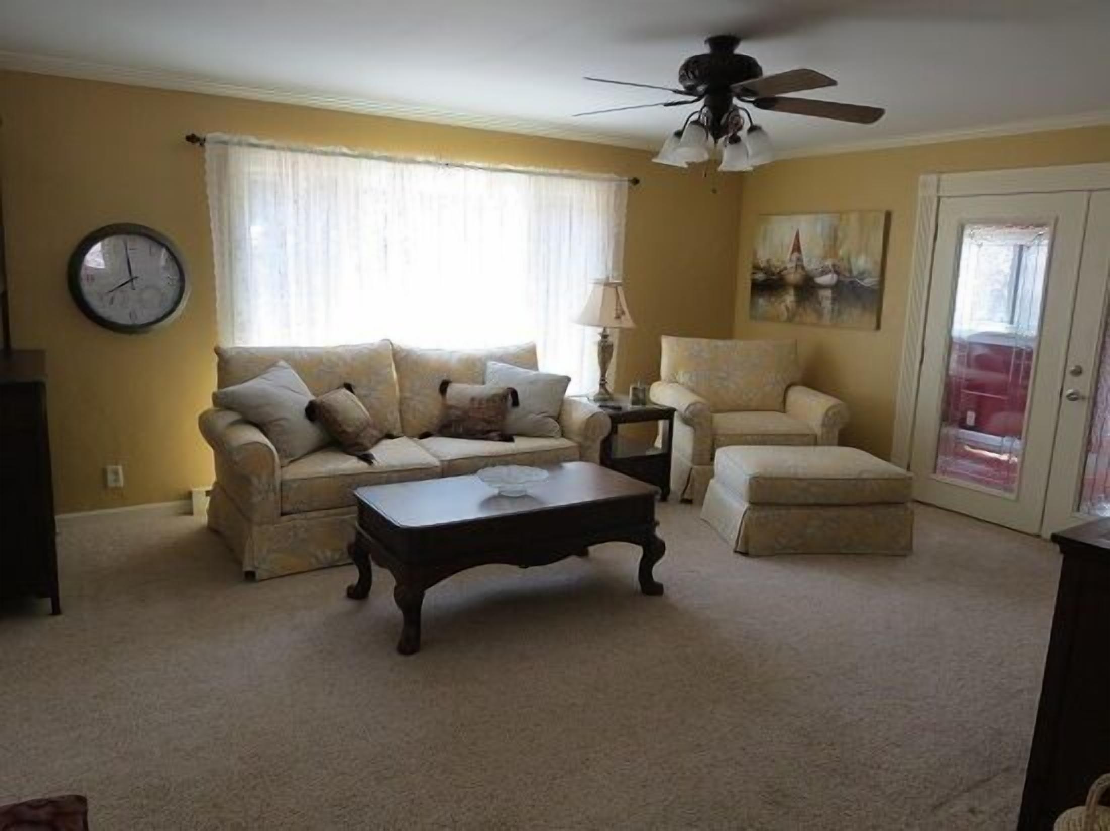 A living room with a beige sofa, a matching armchair, a coffee table, a lamp, a clock, and a ceiling fan.