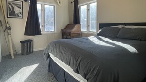 Un dormitorio con una cama grande, dos ventanas con cortinas, un piano y un radiador.