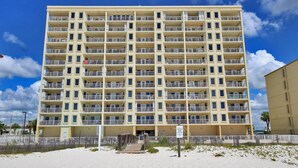 Un hôtel à plusieurs étages doté de balcons, situé en bord de plage, avec une étendue de sable fin devant et un ciel bleu azur.