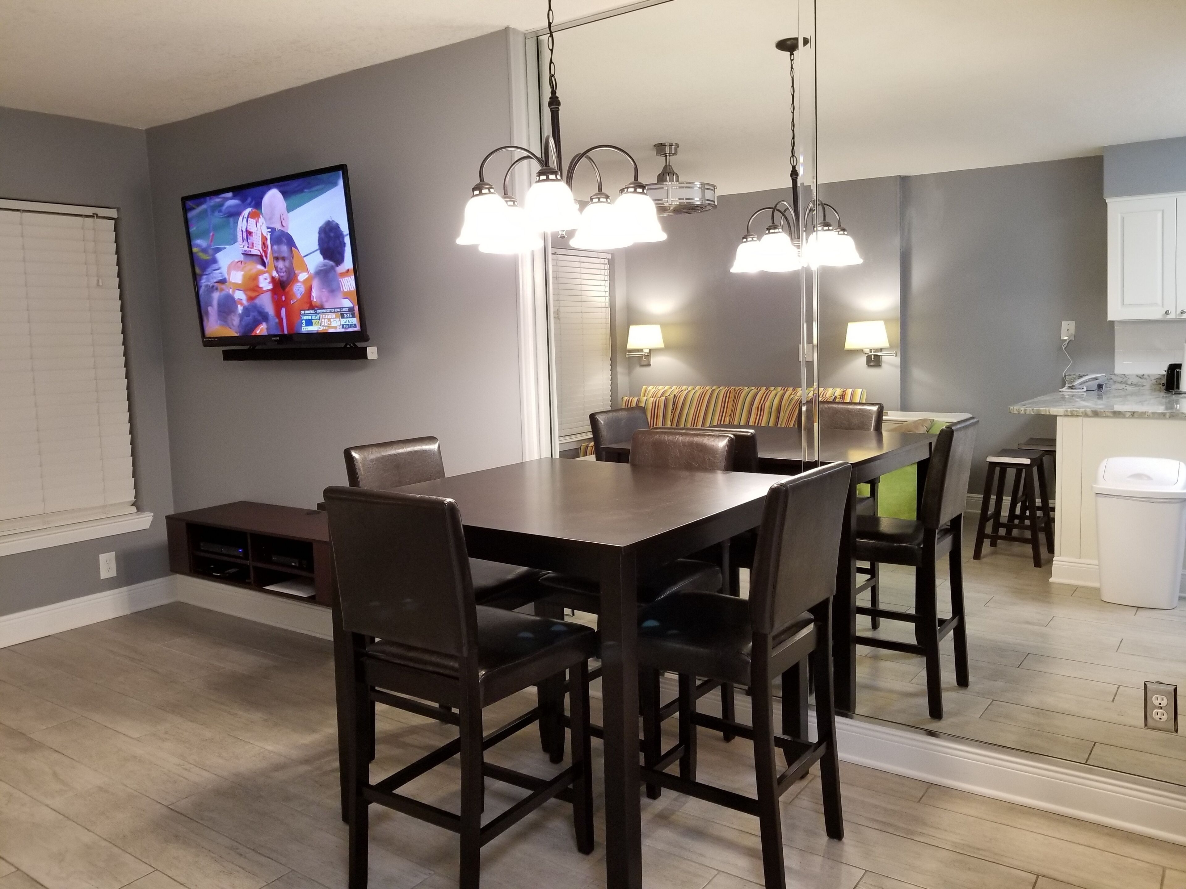 Un espace repas moderne avec une table et des chaises en bois foncé, un téléviseur fixé au mur et des luminaires suspendus au plafond.