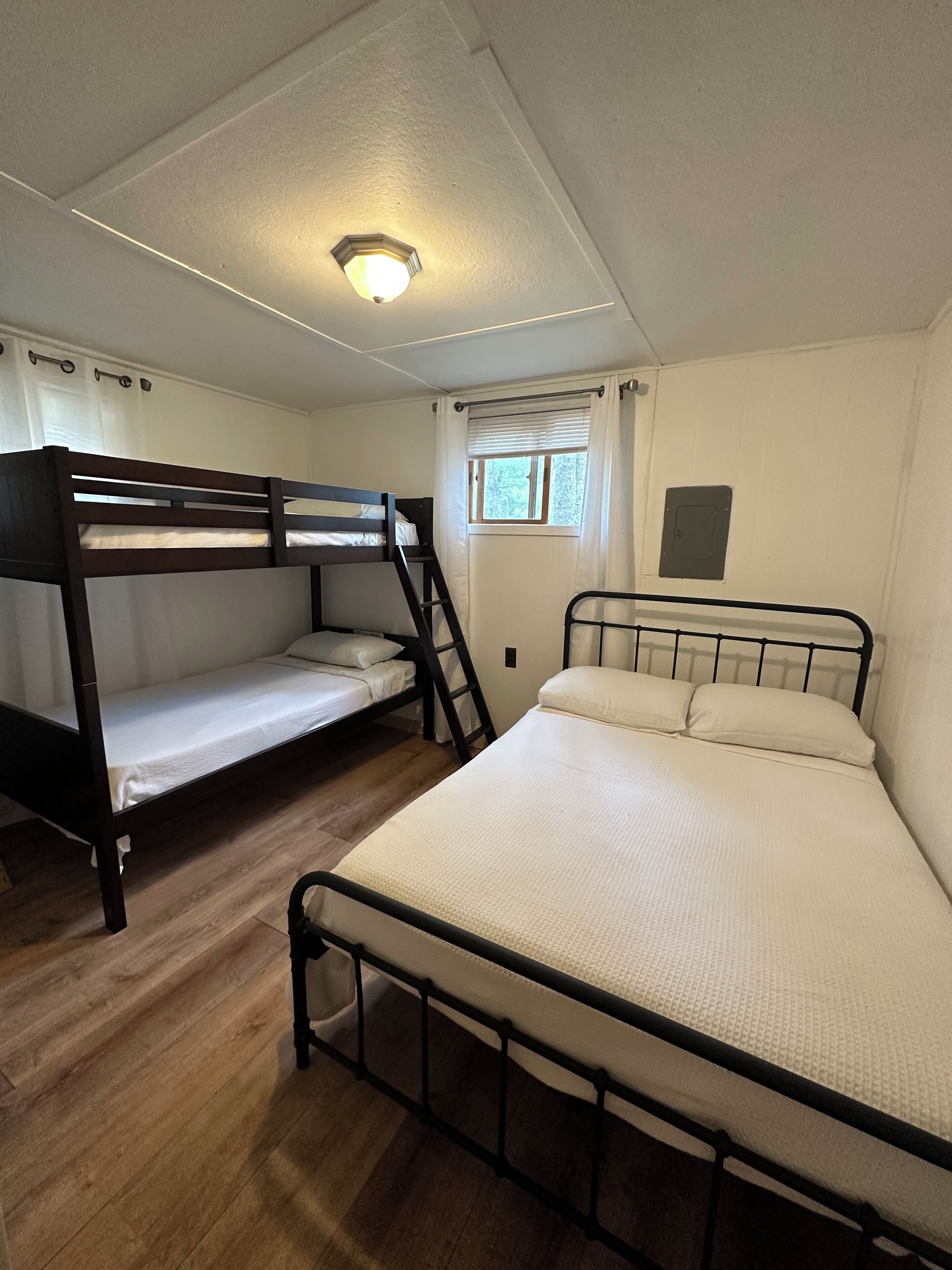 Une chambre de dortoir équipée de deux lits superposés, d’une fenêtre avec stores, d’un luminaire au plafond et d’un petit tableau électrique fixé au mur.