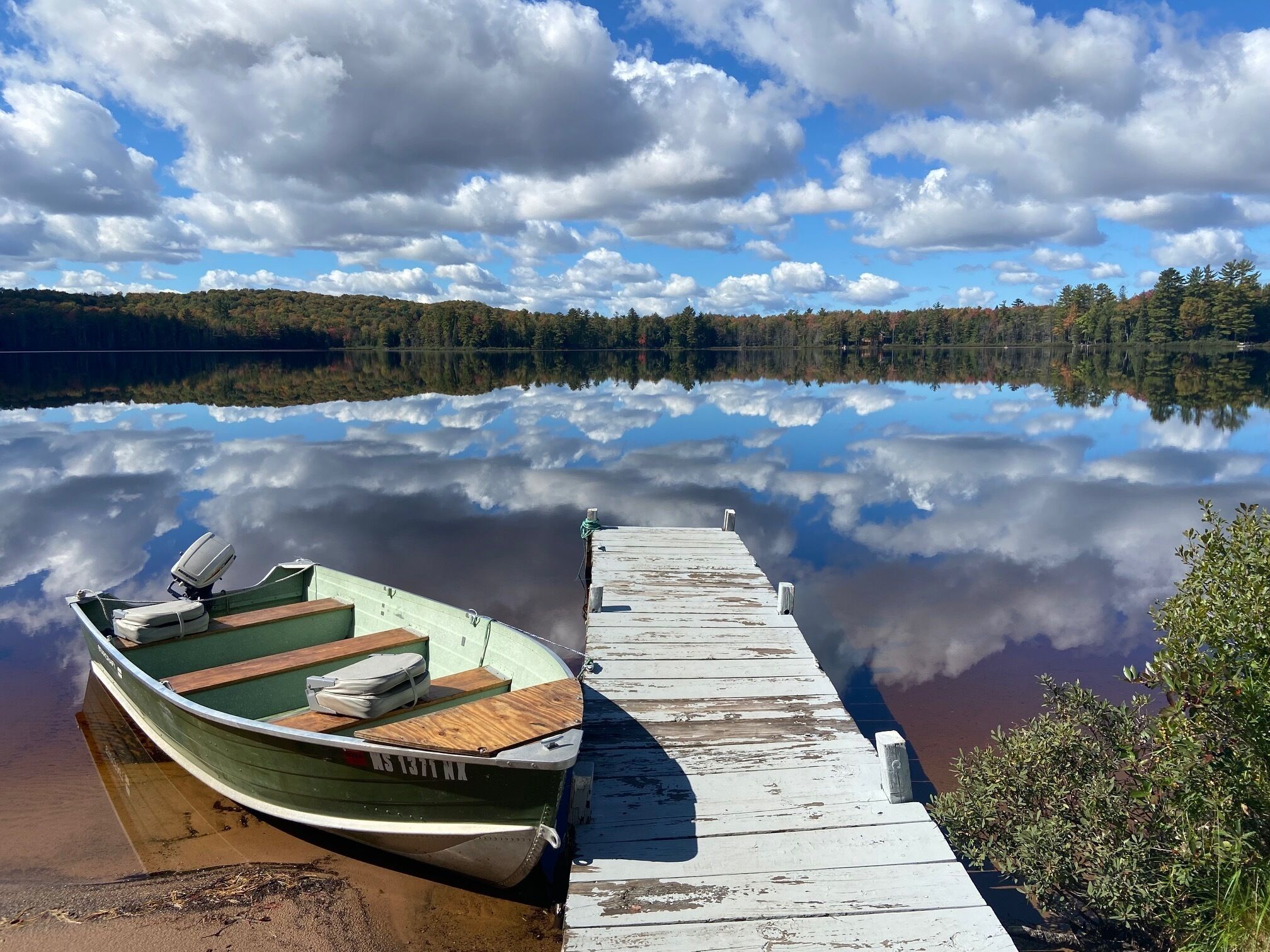 A small green boat docked at a wooden pier extending into a calm lake, surrounded by trees.