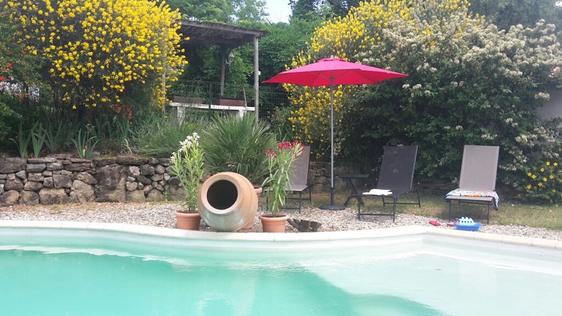 A swimming pool with a stone wall, potted plants, and a red umbrella. There are two lounge chairs and a table with a blanket.
