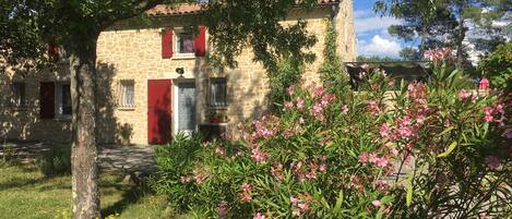 A stone building with red shutters and a garden with pink flowers.