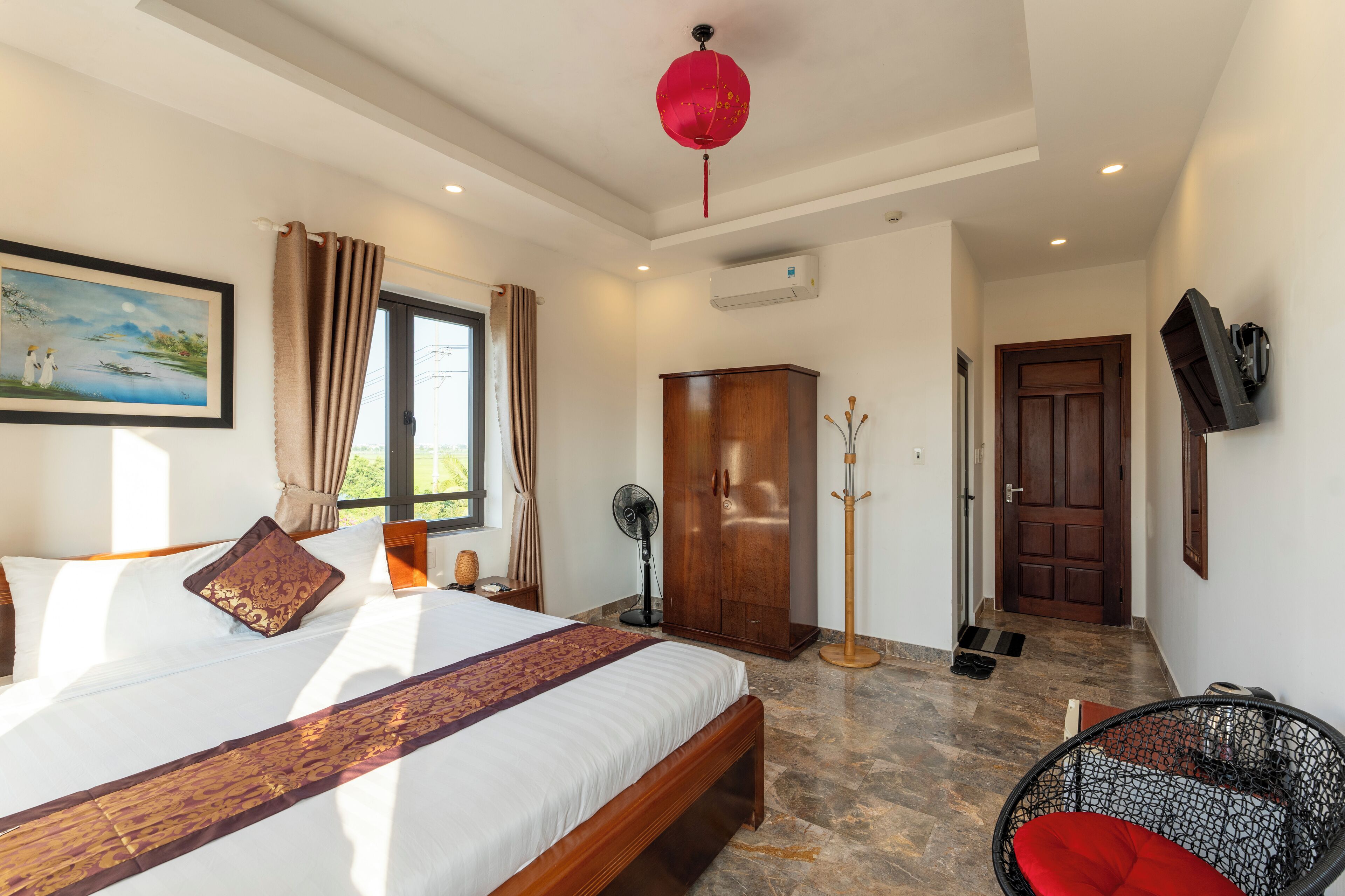 A hotel room with a large bed, a wooden wardrobe, a standing fan, a red lantern, and a television mounted on the wall.