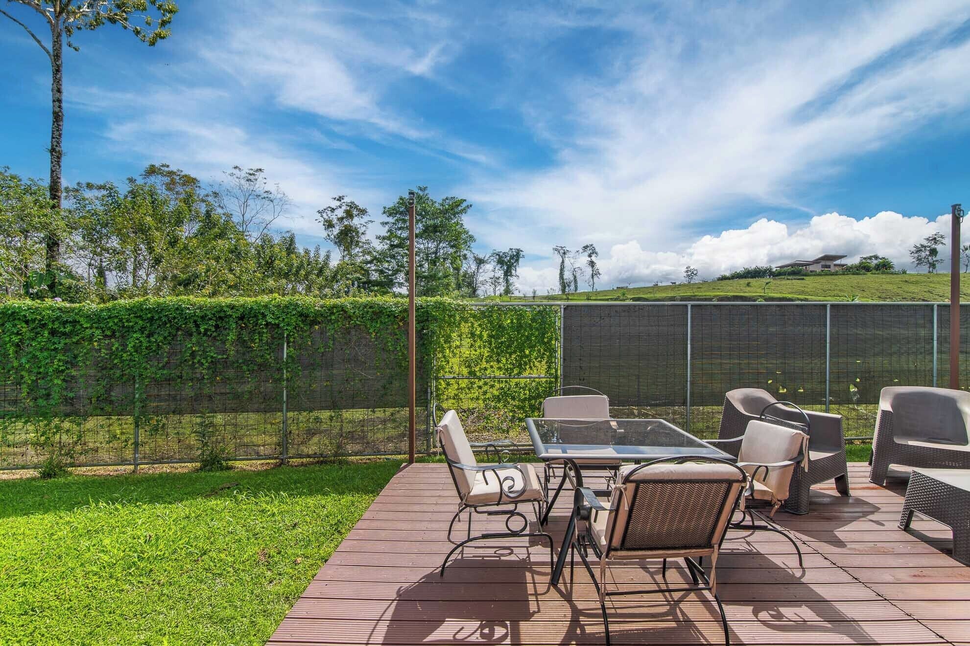 Une terrasse extérieure avec une table et des chaises, entourée d’une plateforme en bois et d’une haie verdoyante.