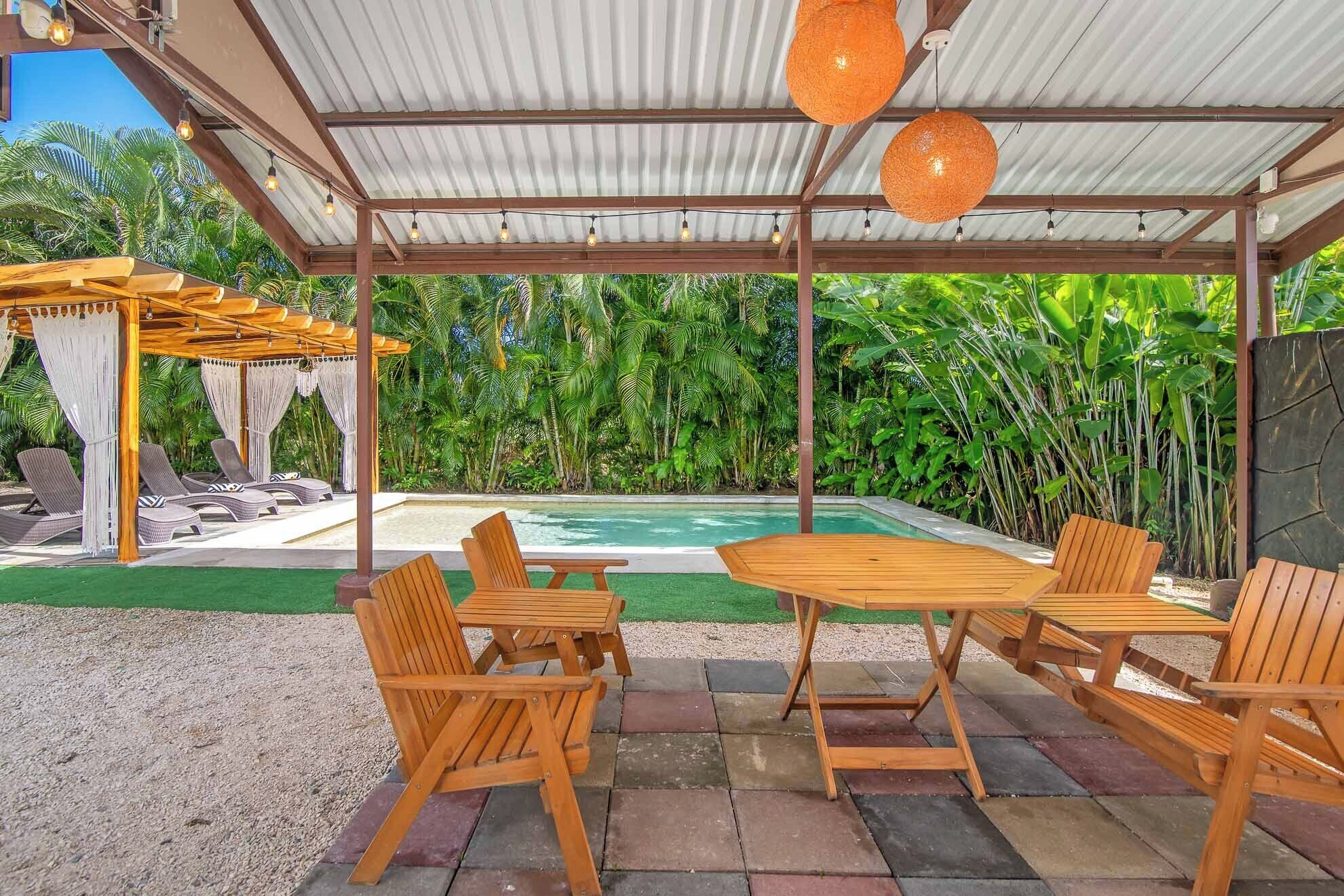 Un espace extérieur abrité comprenant une piscine, des meubles en bois et des plantes tropicales.