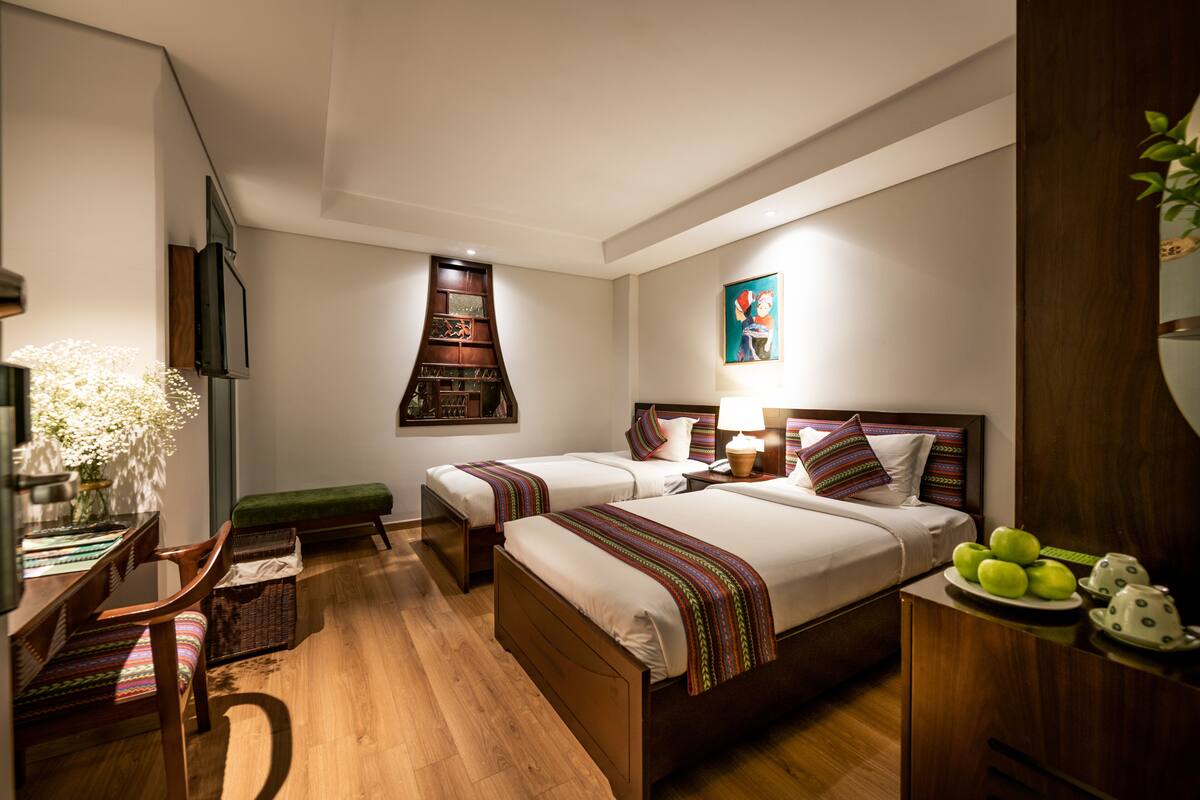 Une chambre d’hôtel avec deux lits, un bureau, une chaise, une télévision, une étagère fixée au mur et un tableau.