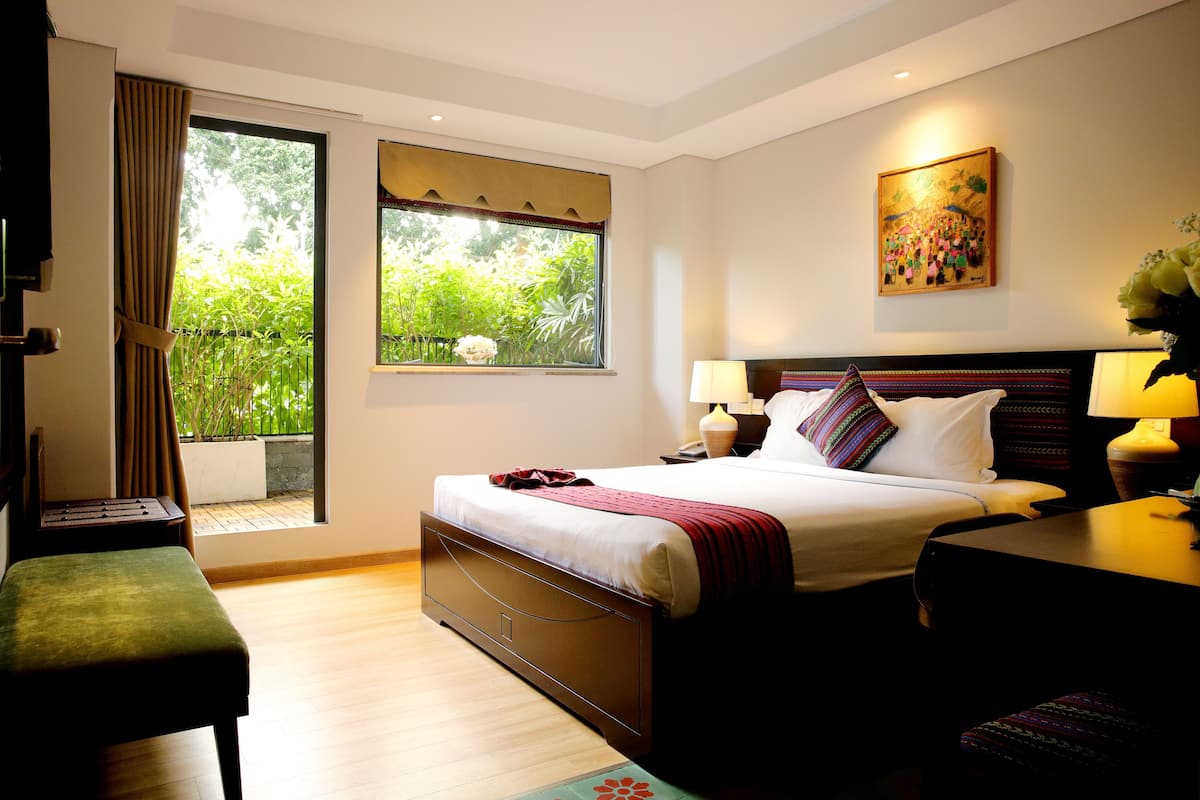 Une chambre d’hôtel comprenant un lit, des tables de chevet, un bureau, une chaise et une fenêtre donnant sur un espace verdoyant.
