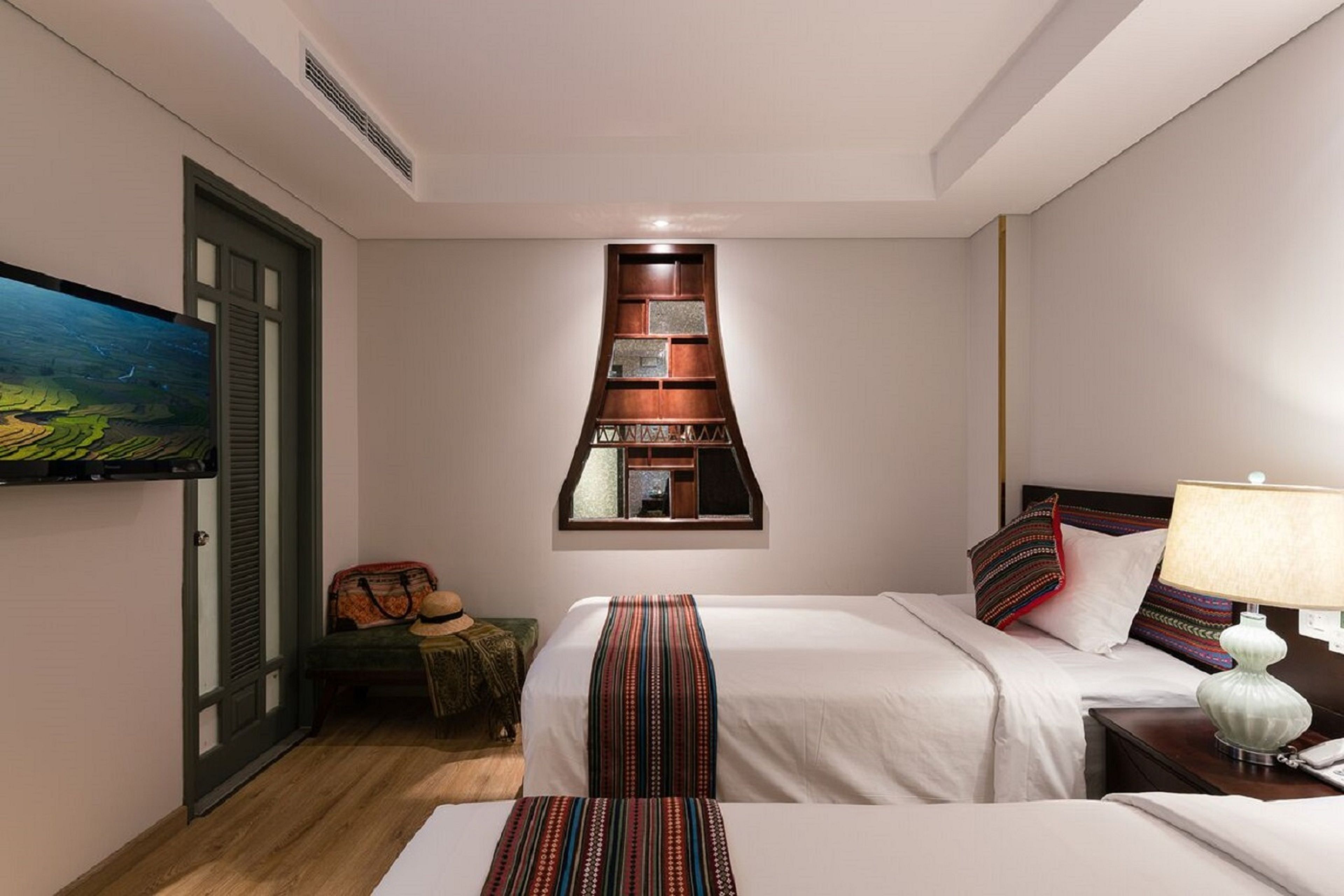 Habitación de hotel con dos camas, cabecero de madera, estante empotrado con objetos decorativos, un cuadro y una lámpara de noche.