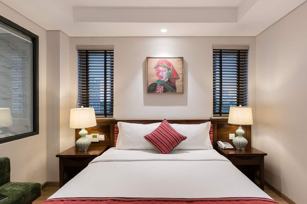 Une chambre d’hôtel avec un grand lit, deux tables de chevet avec des lampes, un fauteuil vert et un tableau au mur.