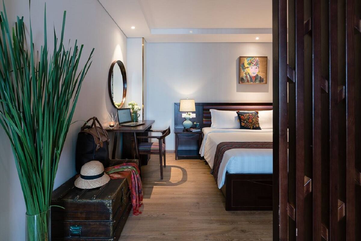 Une chambre d’hôtel avec un lit, une table de chevet, une lampe et un grand miroir. On y trouve une valise et une plante posés sur le sol.