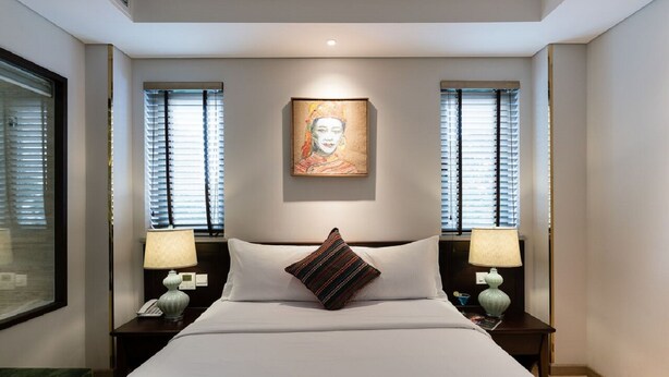 Une chambre d’hôtel avec un grand lit, deux tables de chevet avec des lampes, un tableau au mur et une fenêtre avec des stores.