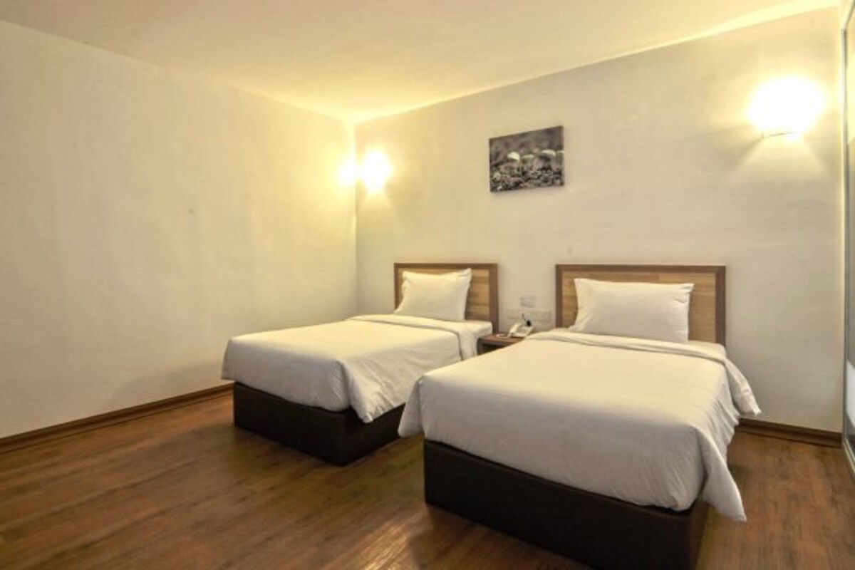 Habitación de hotel con dos camas individuales, suelo de madera y una mesita de noche pequeña con lámpara.