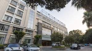 A multi-story building with a modern architectural design, featuring a prominent entrance with glass doors and a sign above. There are several cars parked in front, and the surrounding area includes well-maintained greenery and trees. - Poly Hotel (Guangzhou)