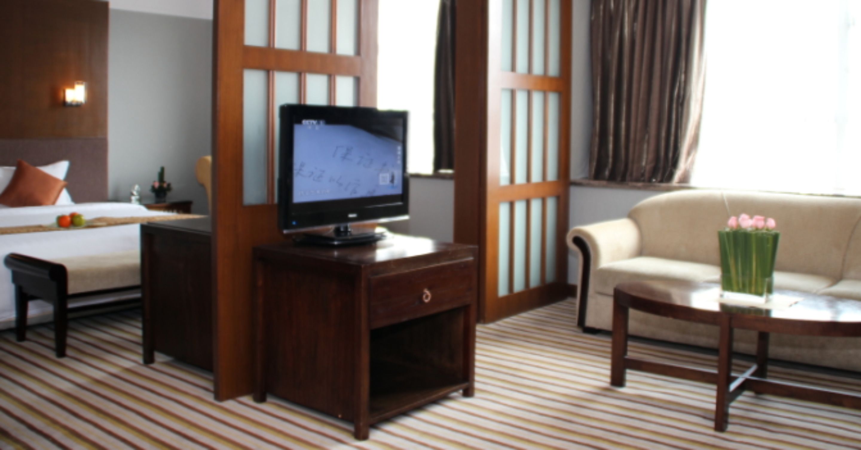 A hotel room with a bed, a TV on a wooden stand, a sofa, and a coffee table with flowers.