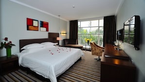 A hotel room with a large bed, a desk, a chair, a TV, and a balcony with a view of greenery. - Poly Hotel (Guangzhou)
