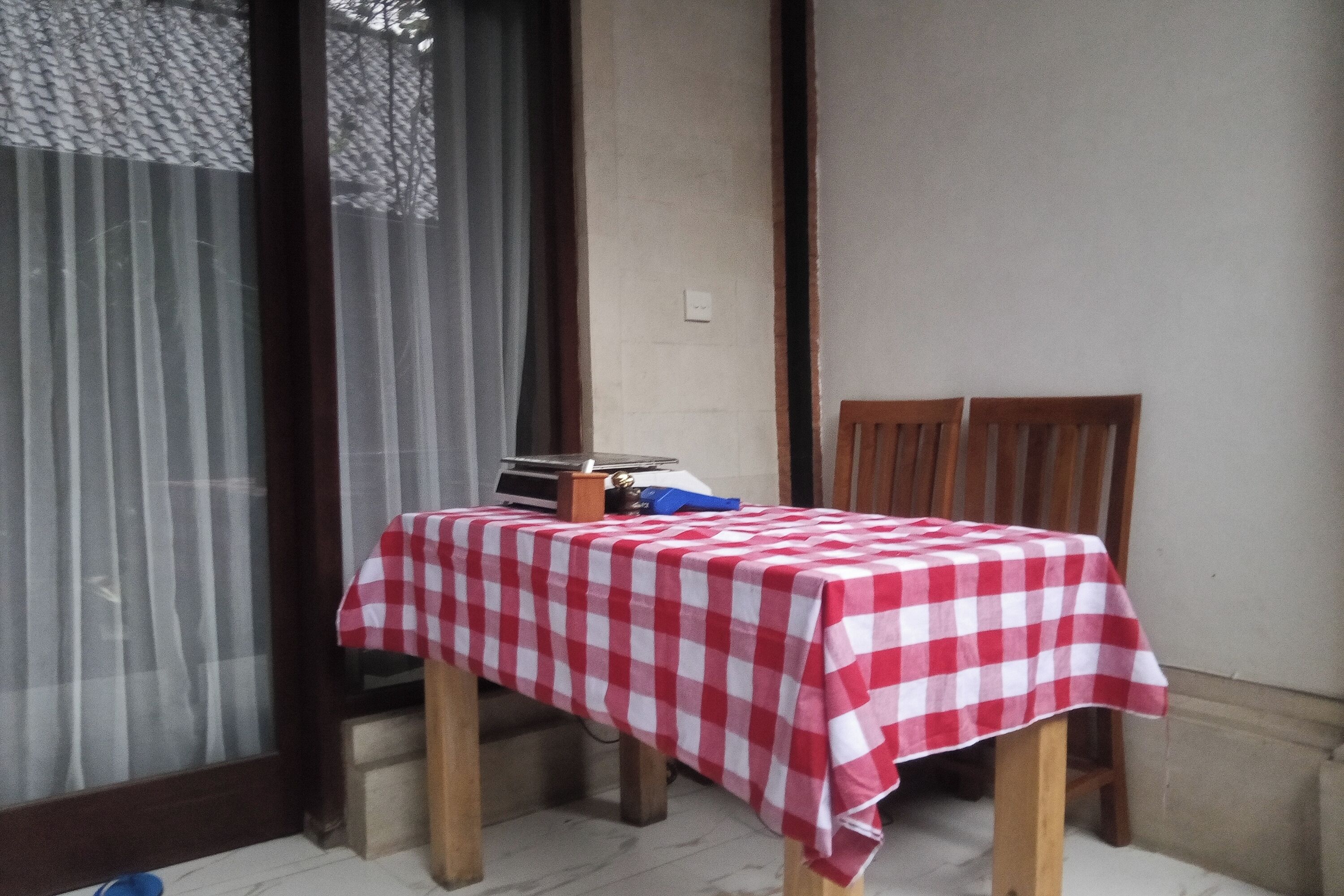 A small room with a table covered by a red and white checkered tablecloth, a chair, and a window with curtains.